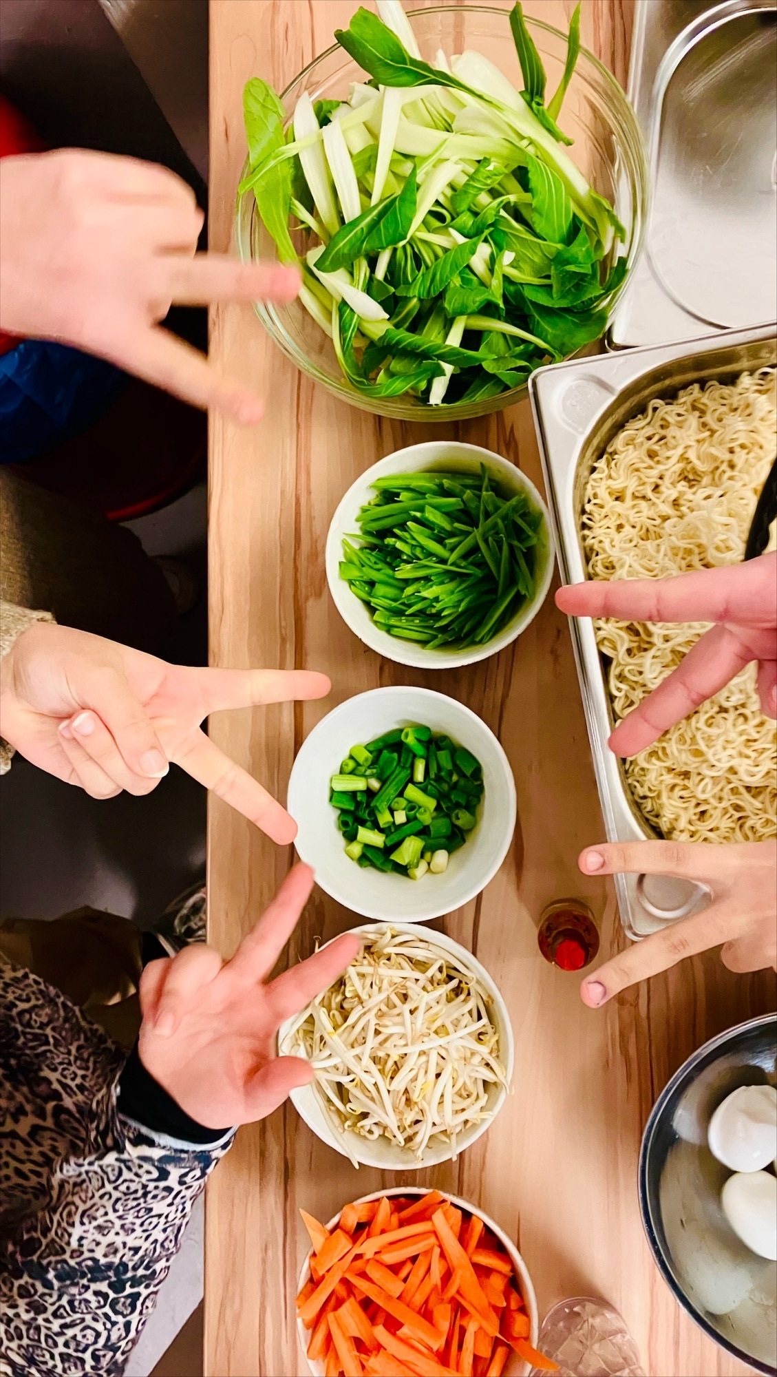 Auf einem Tisch stehen vier runde schalen mit verschiedenem bunten Gemüse. Rechts steht ein großes Gefäß mit Ramennudeln. Der Tisch ist von oben fotografiert. Fünf Hände zeigen das Peace- Zeichen in die Kamera.