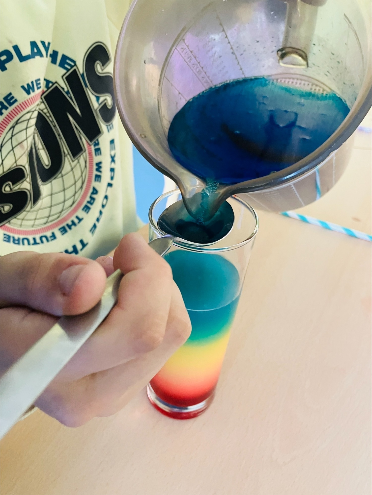 Aus einer Karaffe wird blaue Flüssigkeit in eine Glas geschüttet. In dem Glas befindet sich bereits rote, gelbe und blaugrüne Flüssigkeit.