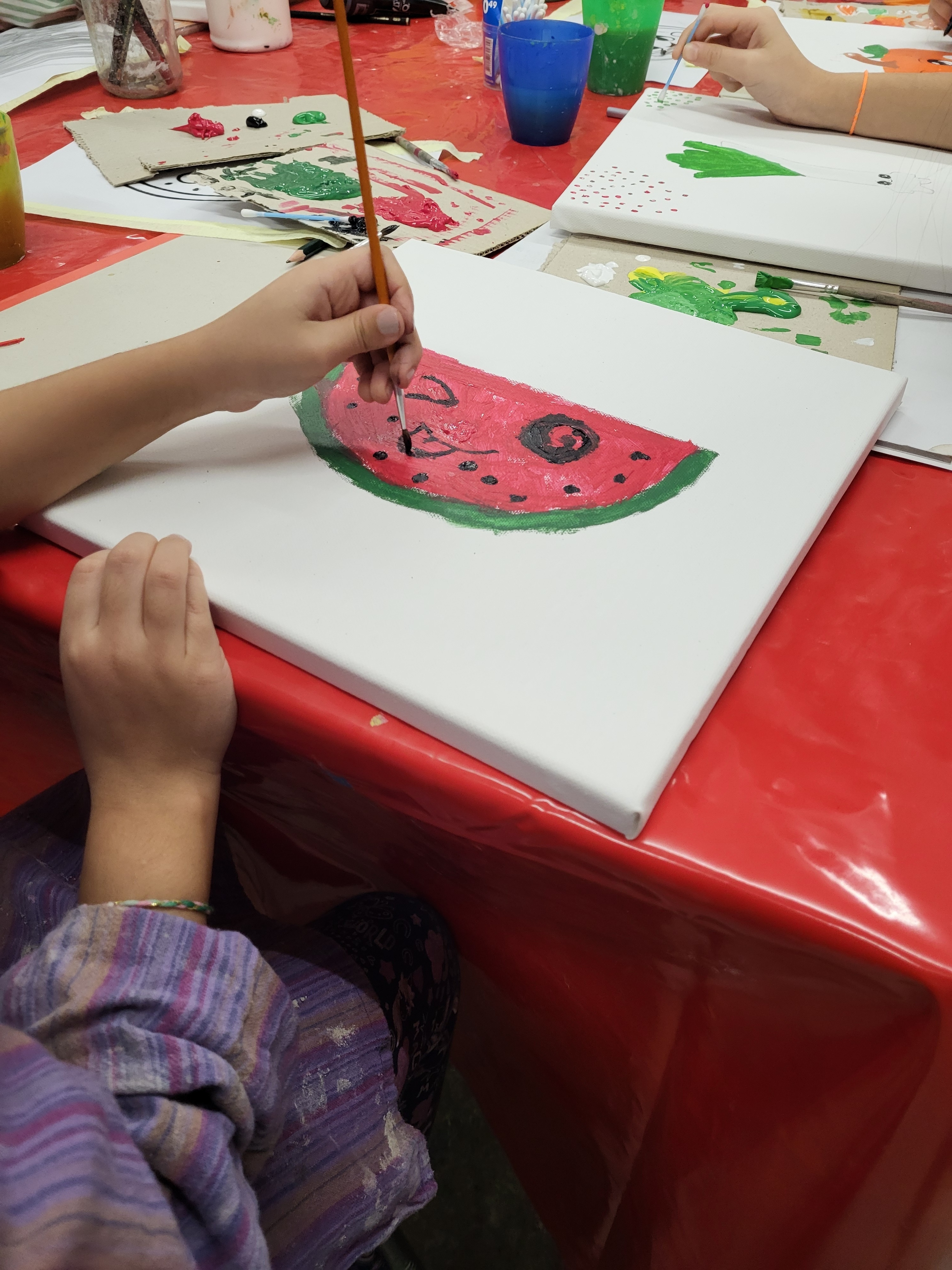 In der Mitte des Bildes liegt eine weiße Leinwand auf einem Tisch, der mit einer roten Basteltischdecke bedeckt ist. Eine Hand hat einen Pinsel in der Hand und mal eine Wassermelone mit Gesicht aus.