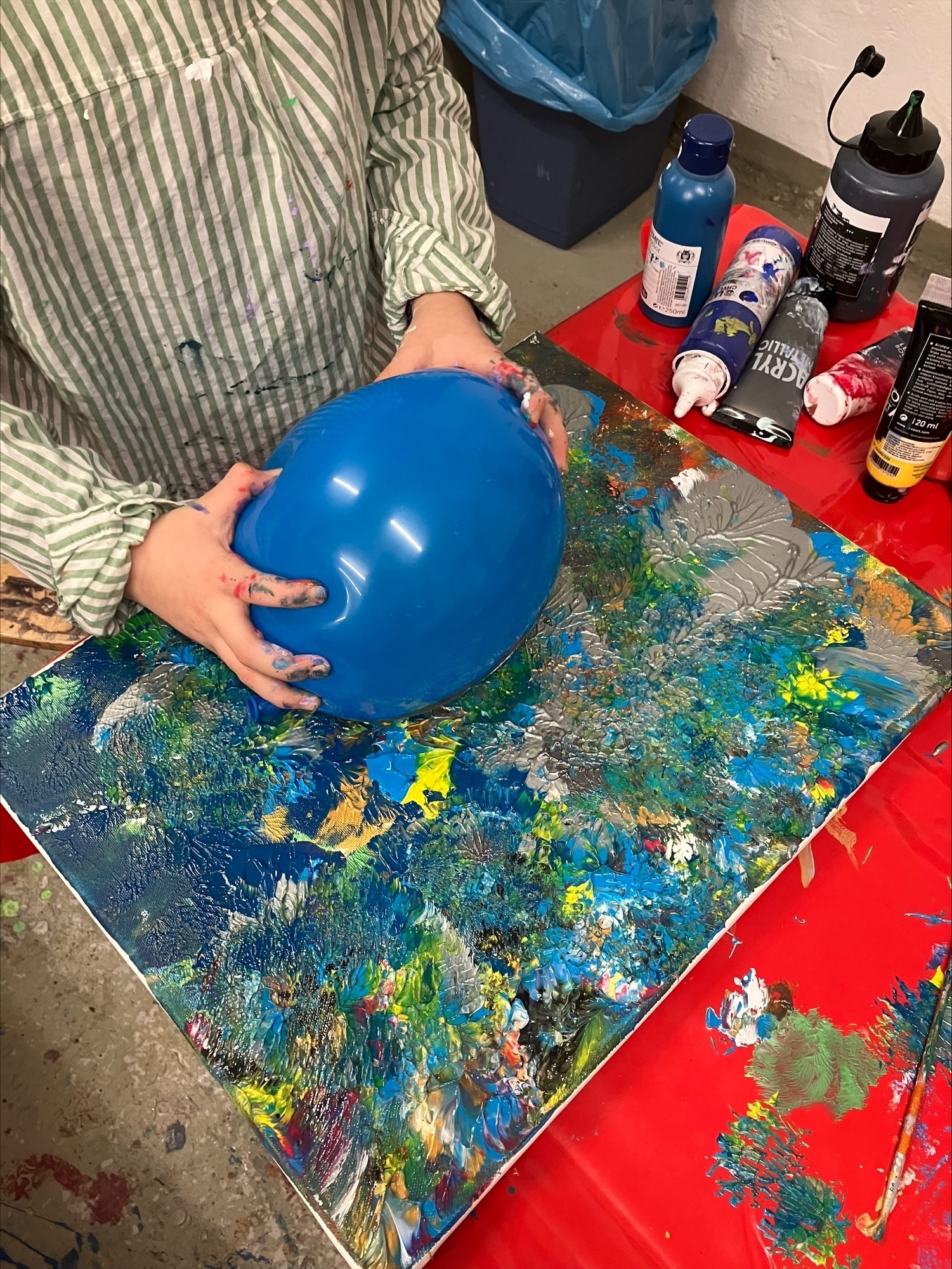Auf dem Bild sieht man eine blauen Luftballon, der mit zwei Händen auf eine Leinwand mit Acryfarbe gedrückt wird.