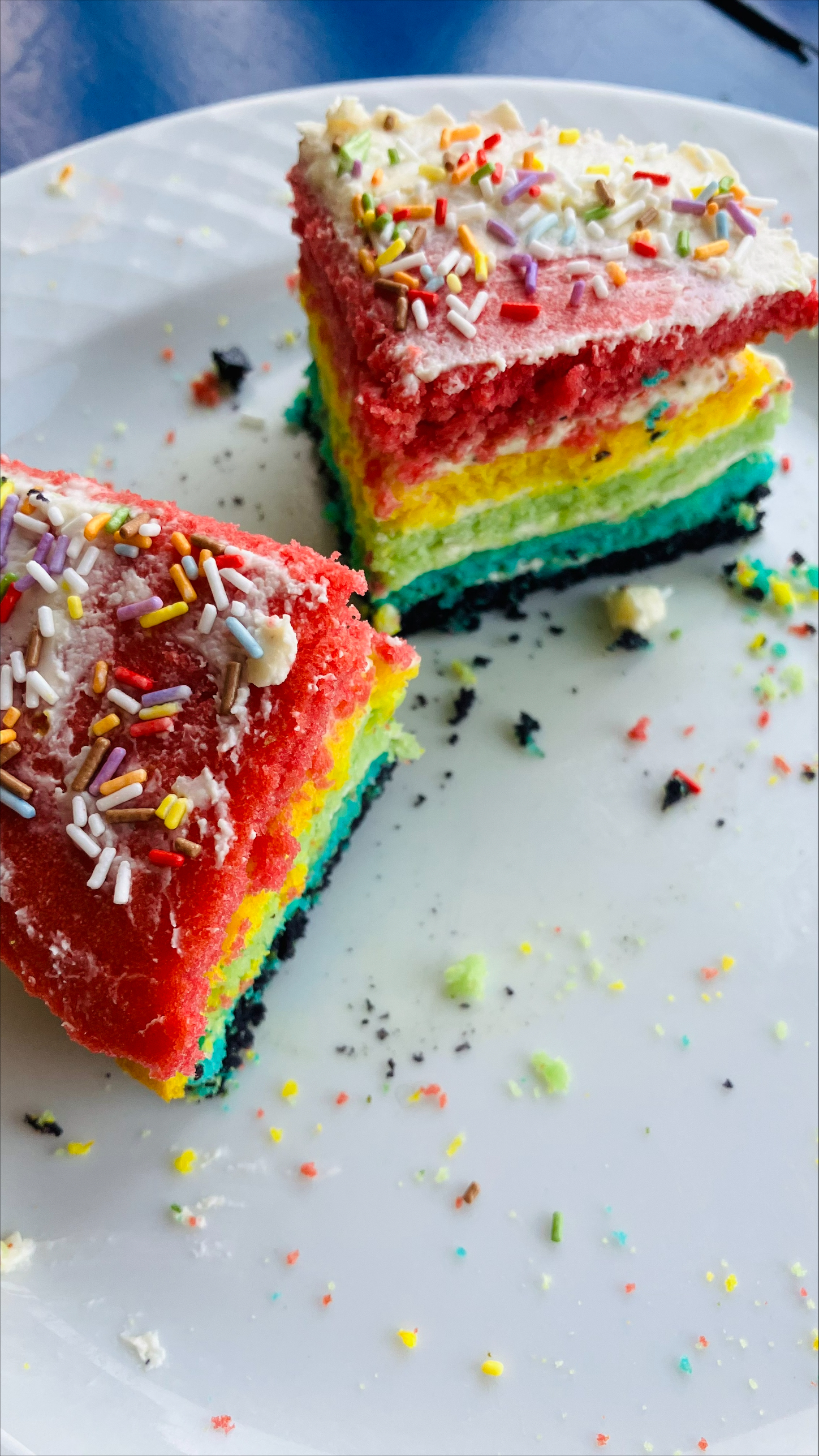 Auf einem weißen Teller stehen zwei Stücke Kuchen. Der Kuchen besteht Schichten in verschiedenen Farben. Oben auf dem Kuchen sind bunte Streusel verteilt.